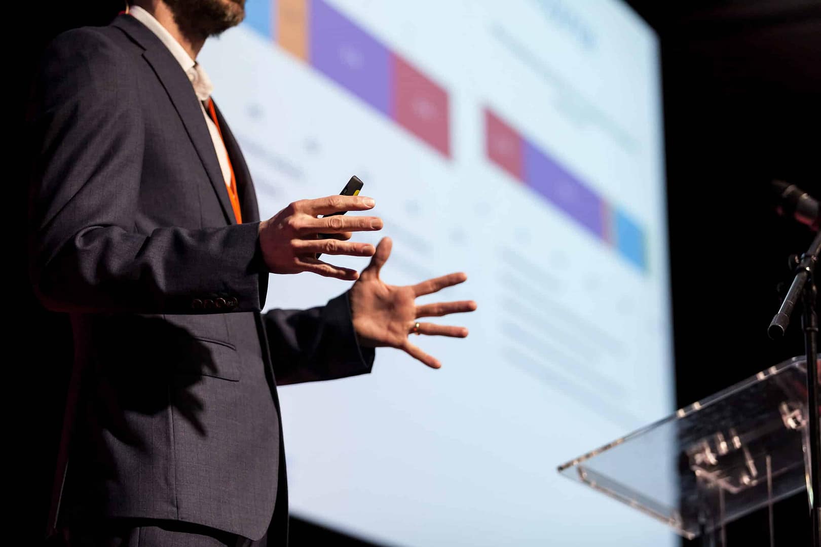 A person in a suit presents data on a screen during a presentation on industry resilience, gesturing with their hands while holding a remote. A microphone and a clear podium are visible in the foreground.