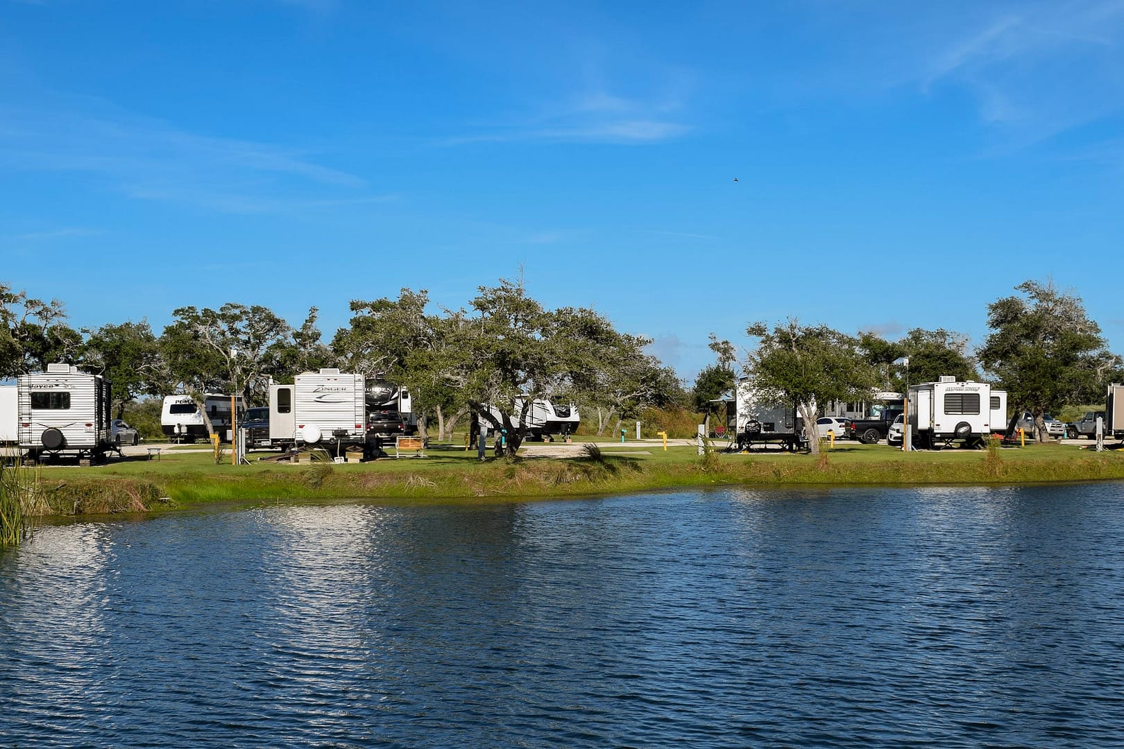 Several recreational vehicles (RVs) parked at a lakeside campsite, with trees and a clear blue sky in the background, reflected on the calm water surface, create an auto draft of serenity.