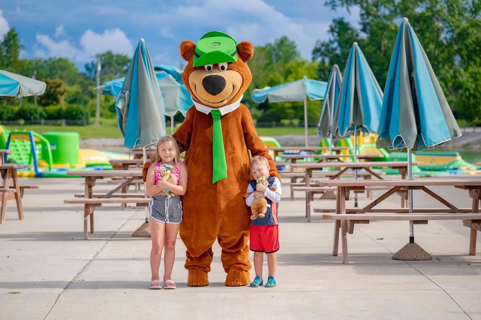 A mascot dressed as a bear standing between two young children, with a water park and auto draft picnic tables in the background.