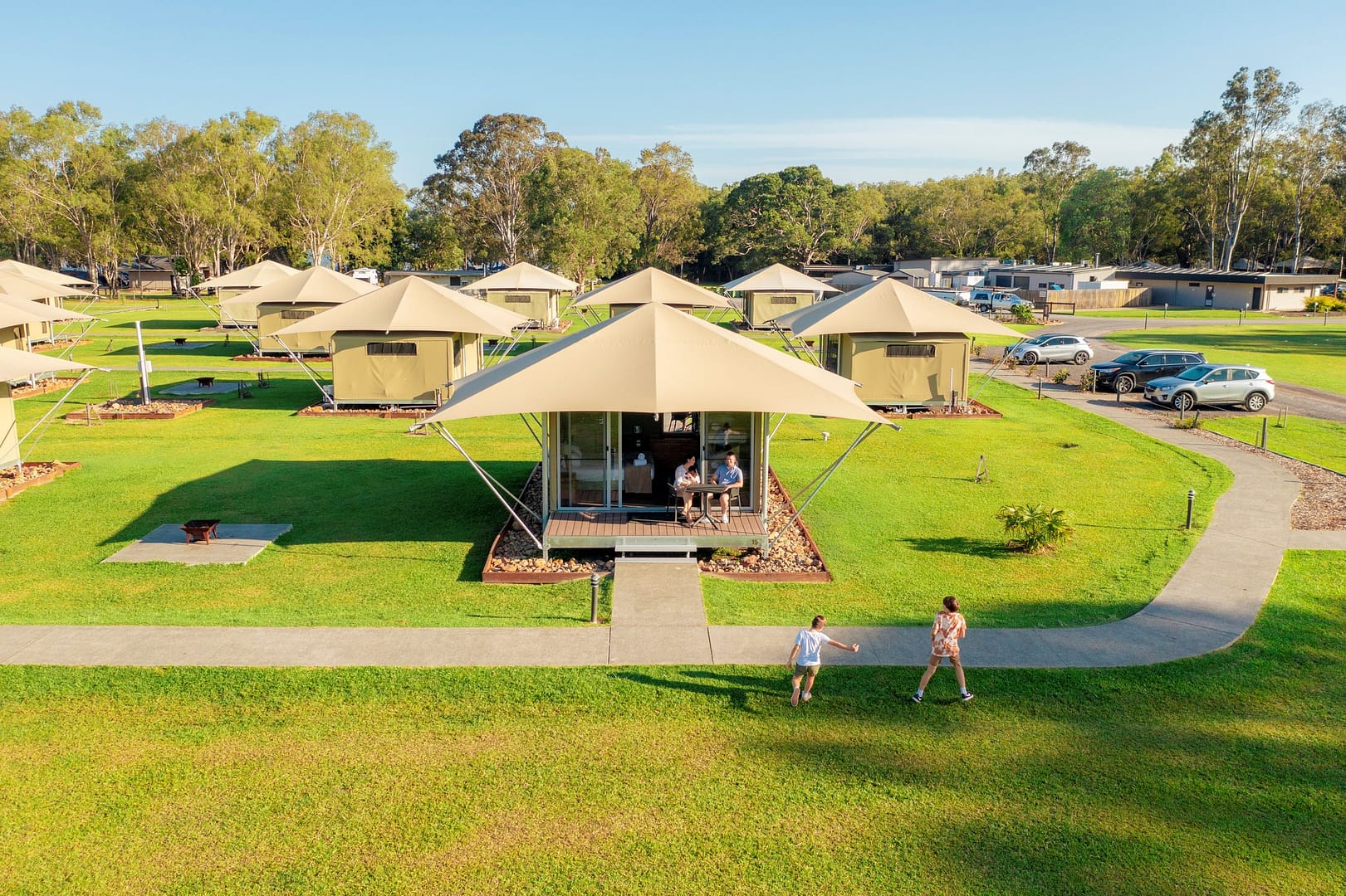 Aerial view of Habitat Noosa campground featuring luxury glamping tents, people walking on green grass, and cars parked nearby.