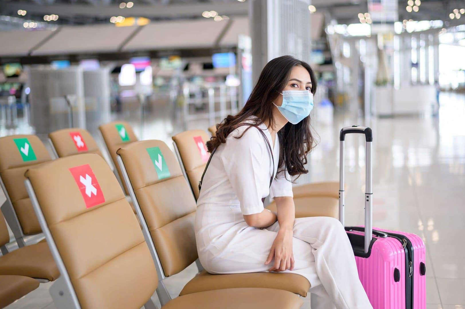 A woman wearing a face mask sits on a bench at an airport, showcasing the ongoing precautions taken in the travel industry.