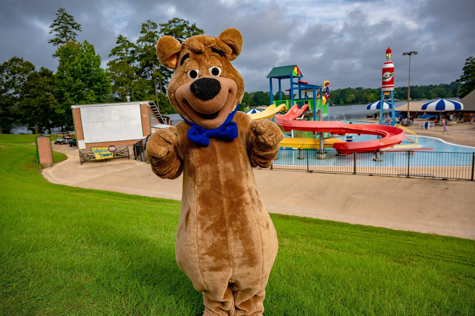Person in a bear costume with a blue bow tie posing in front of a water park with slides, trees, and a lake in the background, seamlessly blending into the lively auto draft of fun and adventure.