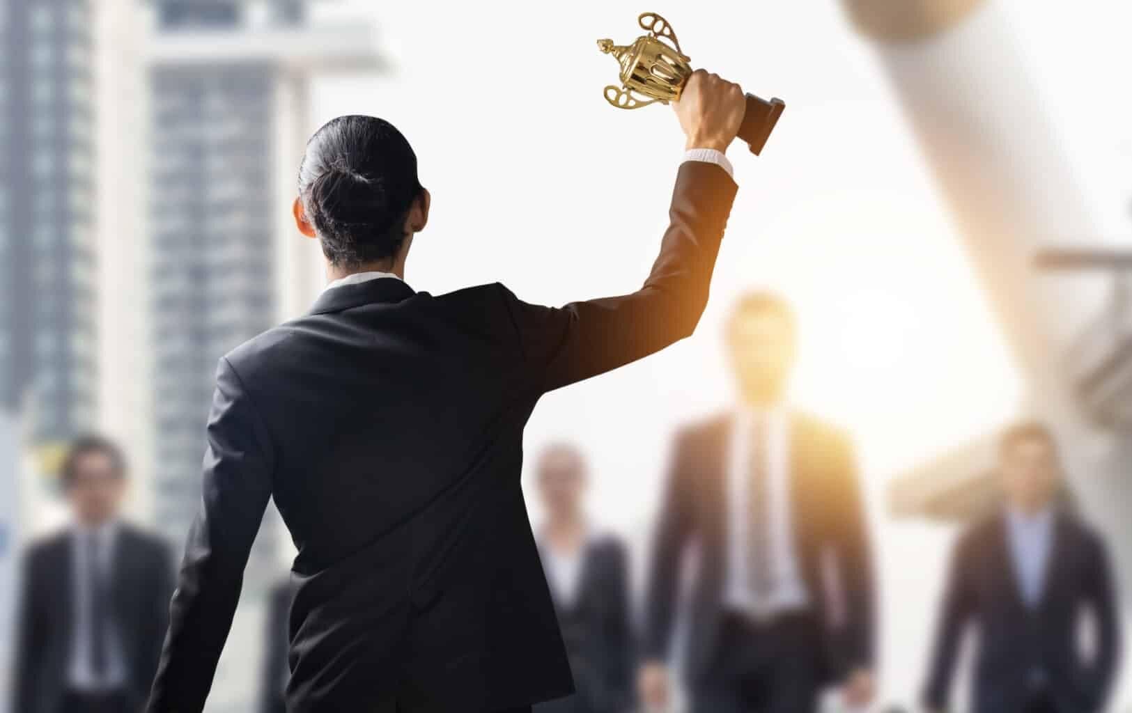 A person wearing a suit holds up a trophy with one hand, while several other individuals in business attire walk towards the person from the background, reminiscent of a celebratory scene at one of Boston's top corporate award events.