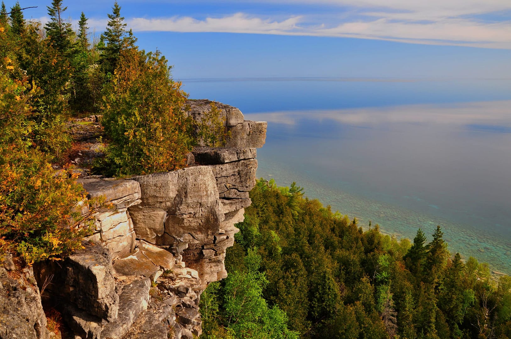 A cliff adorned with greenery overlooking a calm body of water under a partly cloudy sky. Auto Draft