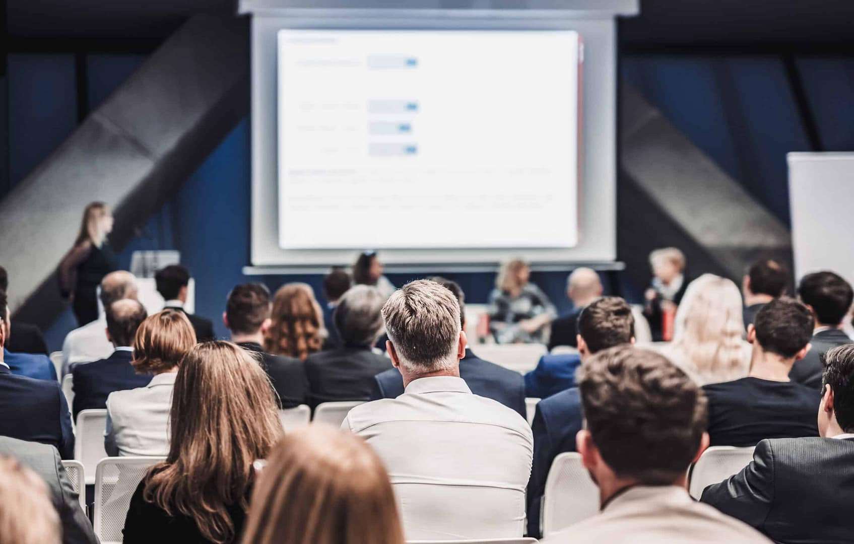Rear view of business professionals attending a Trade Show with presenters onstage discussing slides.