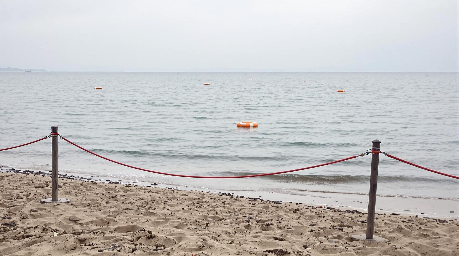 A deserted lakeside with roped-off shoreline and orange float barriers indicating a closed swimming area, captured on an overcast day with gentle ripples on the water and an empty beach.
