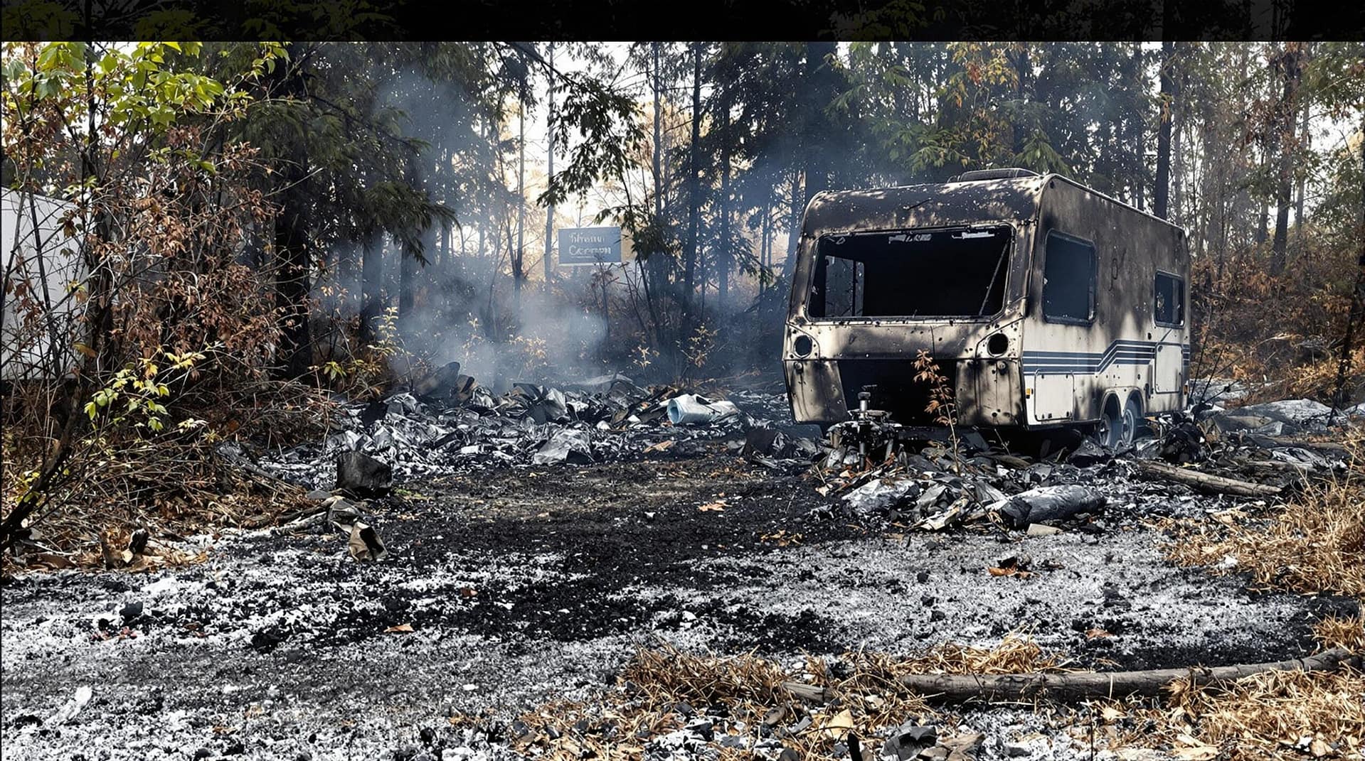 Scorched campground with burnt-out camper and charred grass, smoke lingering in the air.