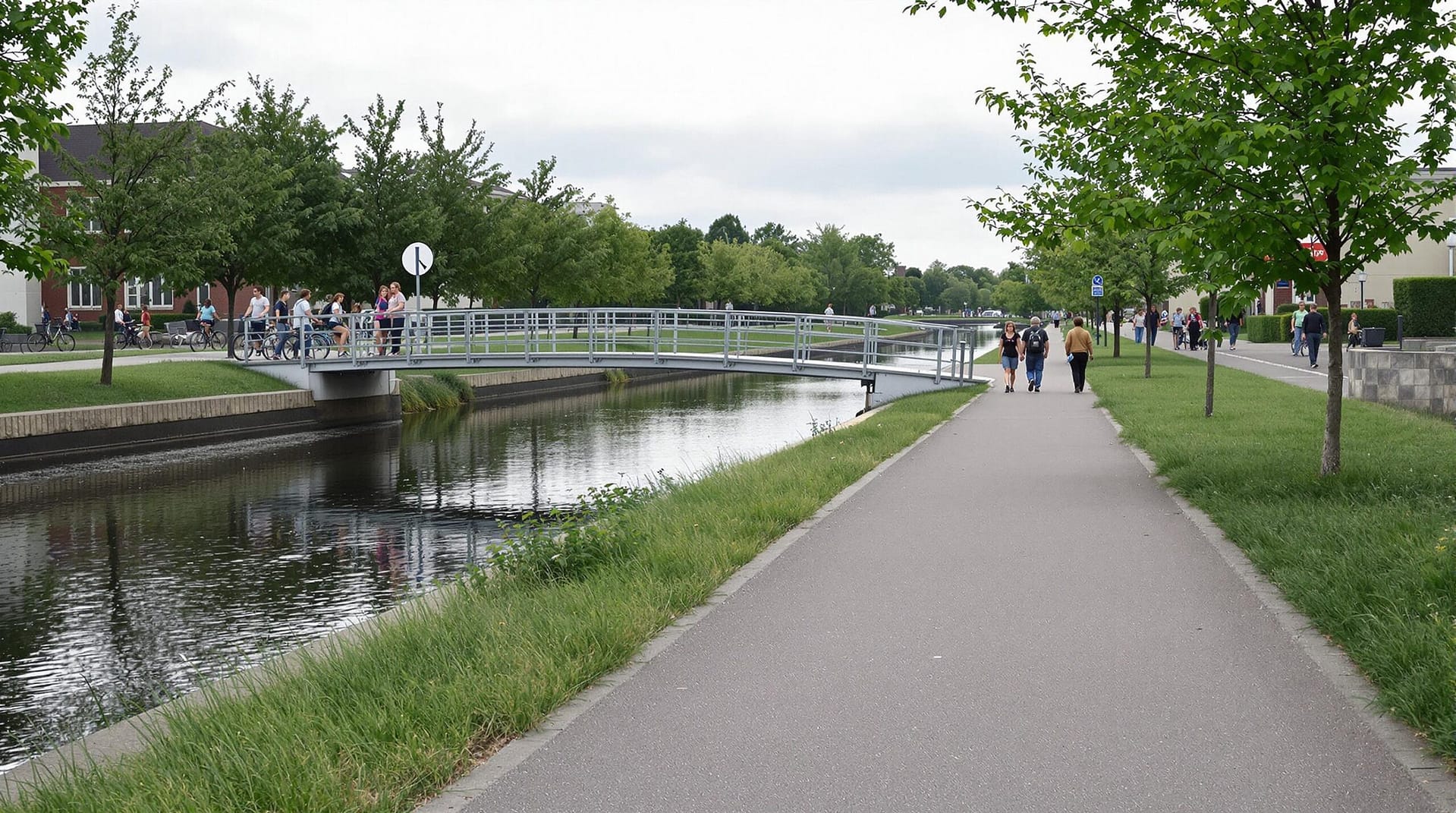 A scenic canal-side walking and biking path with a small pedestrian bridge and several people enjoying the trail on an overcast day, surrounded by greenery and neutral buildings.