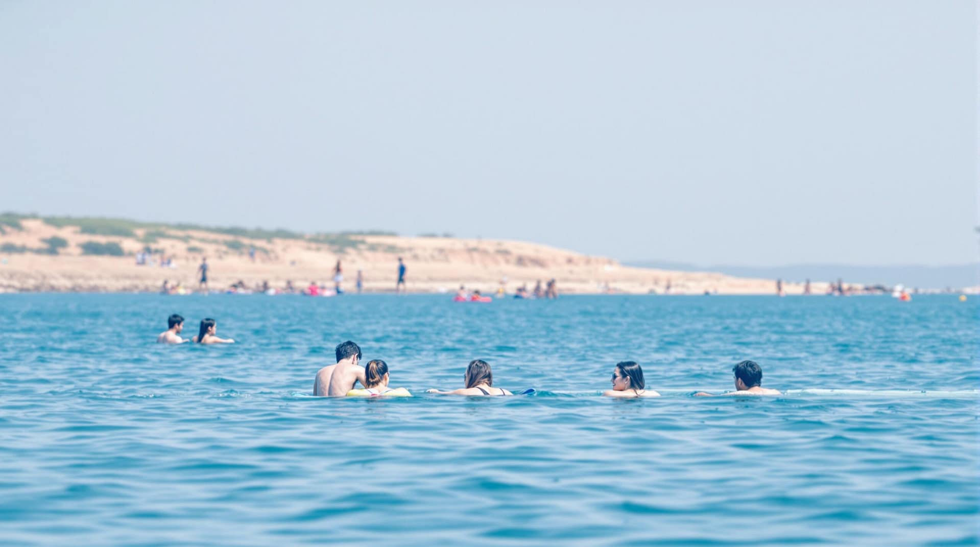 A group of people enjoying recreational activities near a tranquil waterfront, with calm blue water and a sunny, clear sky in the background.