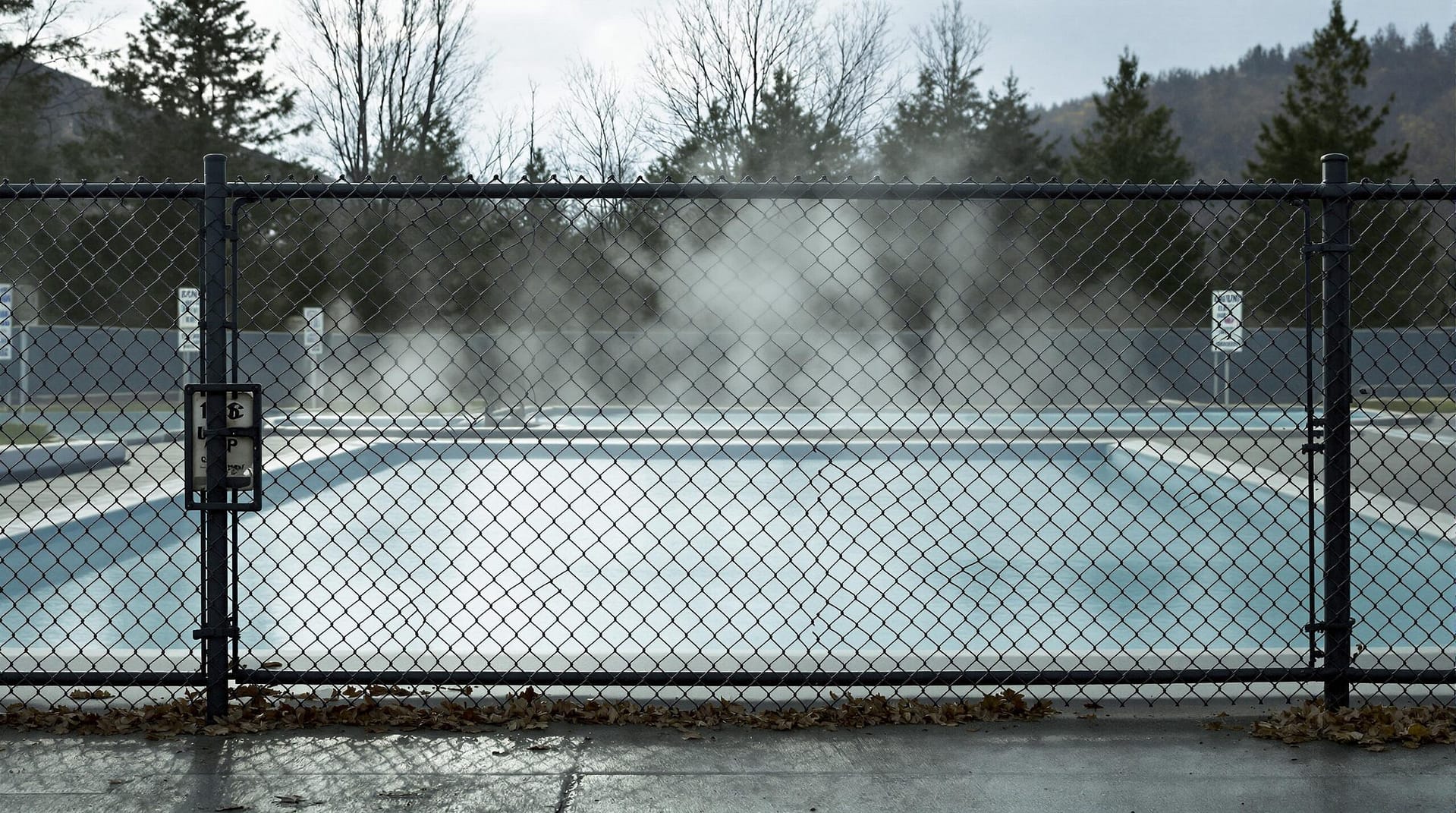 An empty outdoor hot springs pool behind a locked chain-link fence, with steam rising gently from the water under muted daylight, creating a quiet and subdued atmosphere.