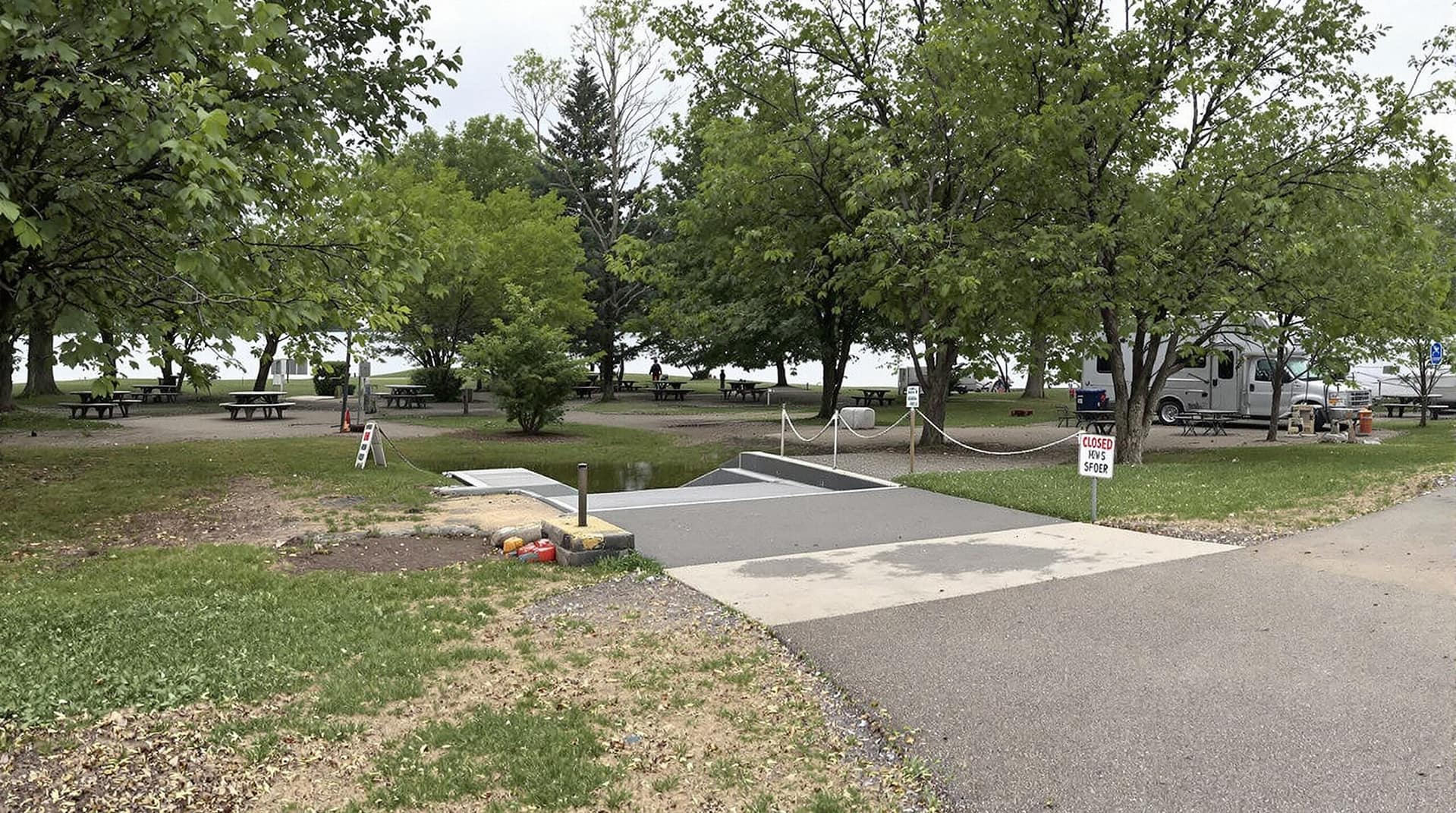 A quiet riverside campground with a small boat ramp, empty picnic tables, and sparse greenery under a cloudy sky, indicating minimal activity.