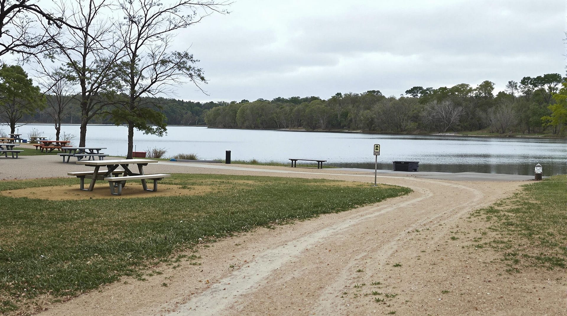 A serene riverbank recreation area with a dirt path leading to a calm river, sparse picnic tables, and short grass under an overcast sky, with trees lining the river in the background.