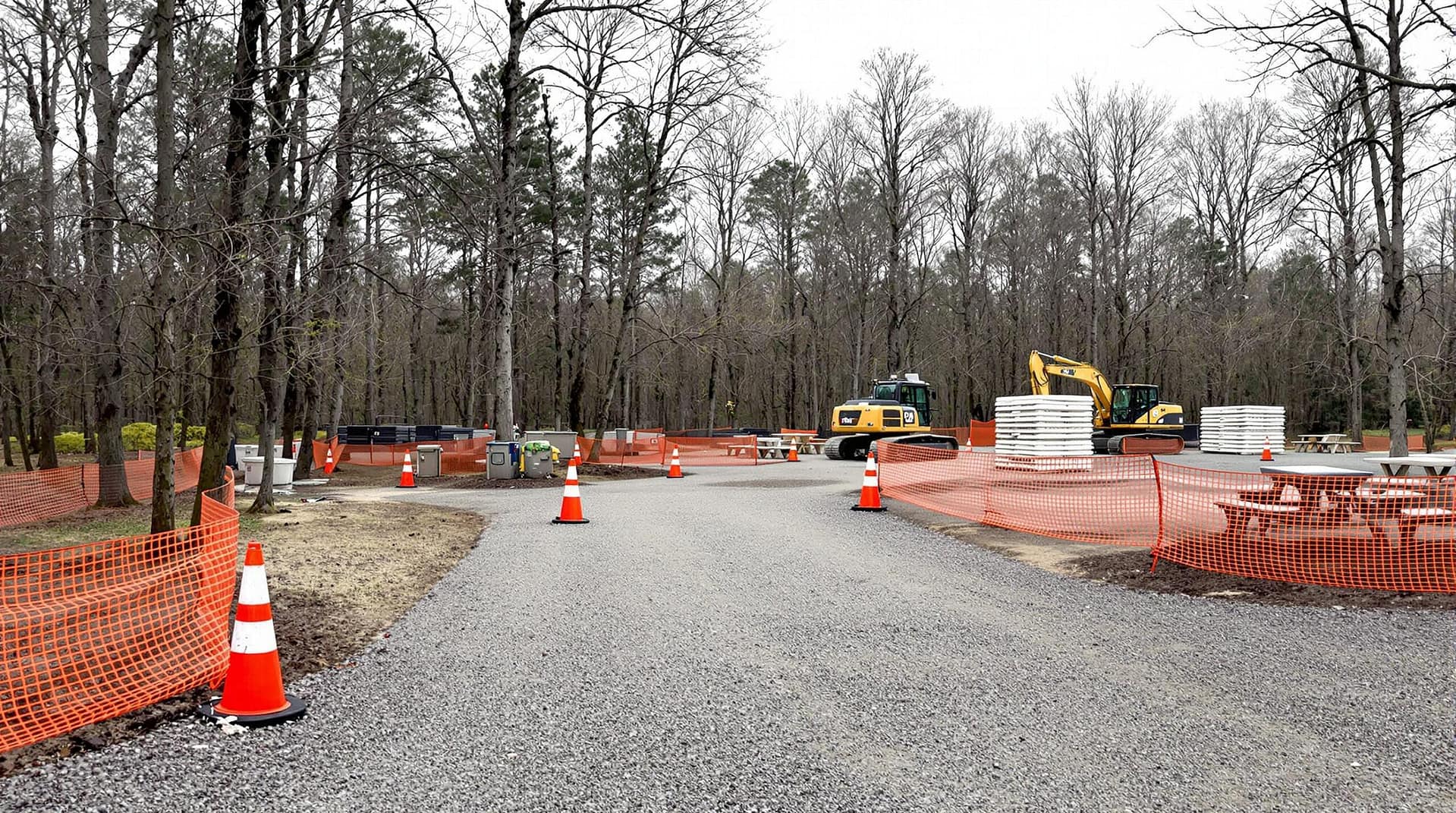 Renovation scene at a closed campground loop with empty RV pads, orange construction fencing, a yellow excavator, new picnic tables, and leafless trees in a generic forested area
