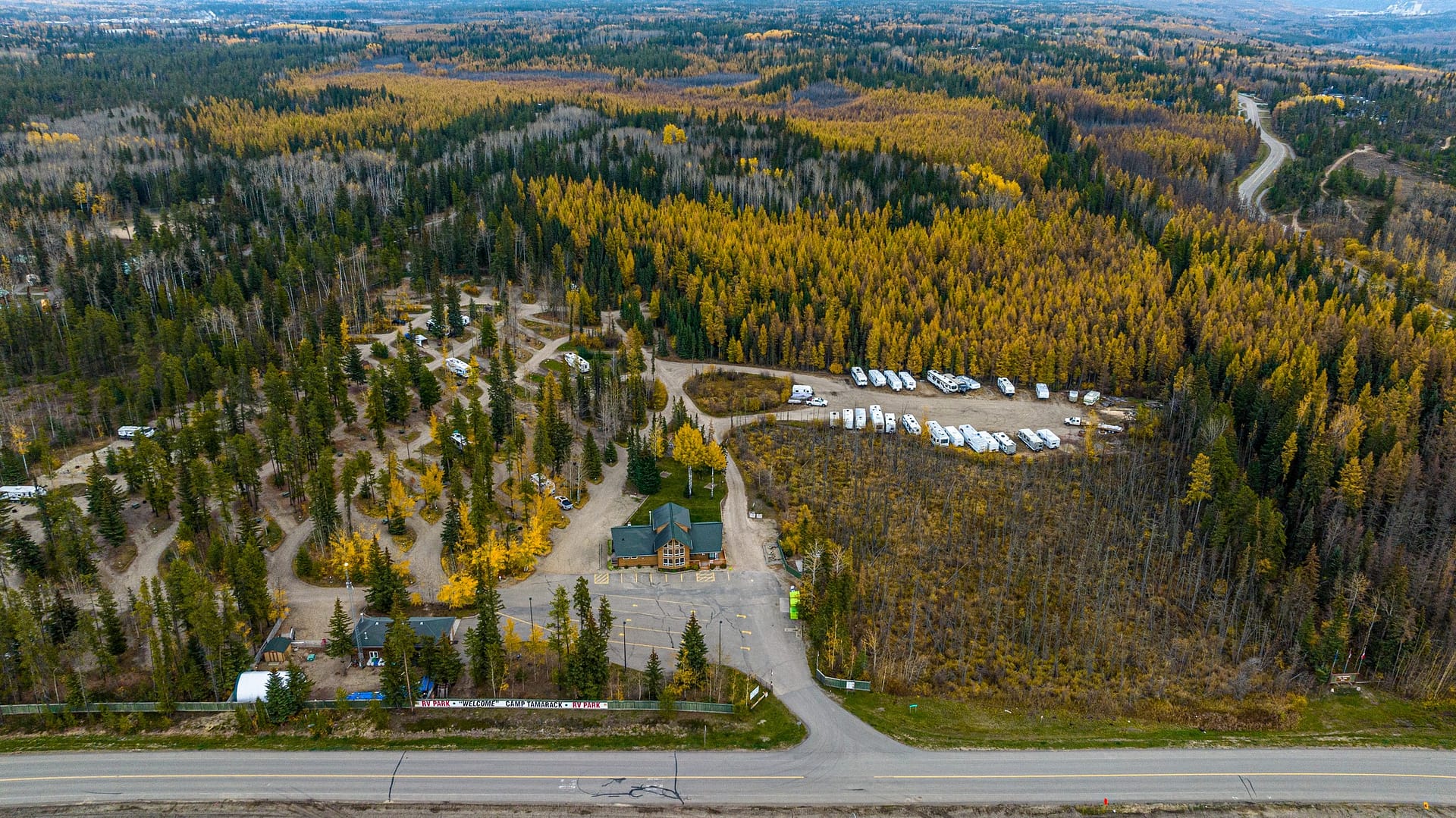 Aerial view of the Grande Prairie area with a mix of evergreen and autumn-colored trees, a small cluster of buildings at Camp Tamarack RV Park, and a car park surrounded by winding roads
