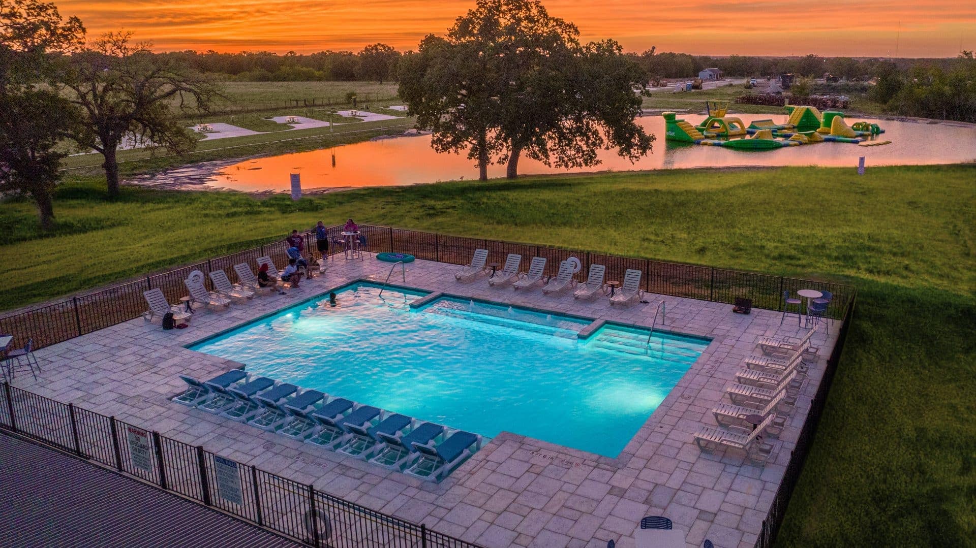 A swimming pool is surrounded by lounge chairs near a lake with an inflatable water park. A group of people is gathered on one side of the pool, chatting and enjoying the evening. The sky, bathed in hues of orange and pink from the setting sun, seems almost like an auto draft painting.