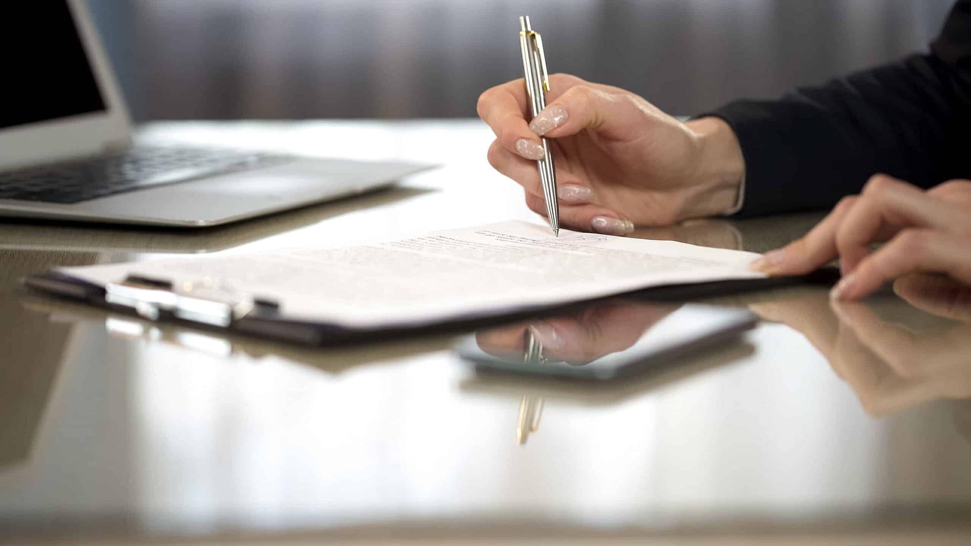 A person reviews regulations at a desk with a laptop open nearby, preparing to write with a pen in hand.