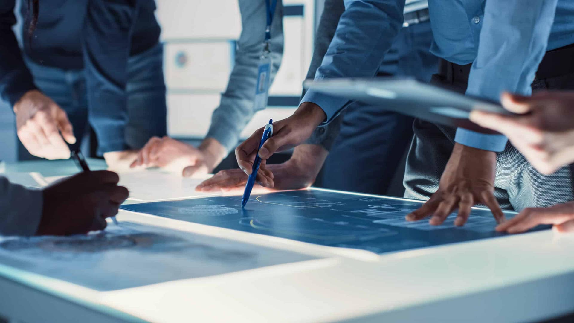 A group of professionals analyzing and discussing digital data on an illuminated table in a modern office setting overlooking a mini golf course.