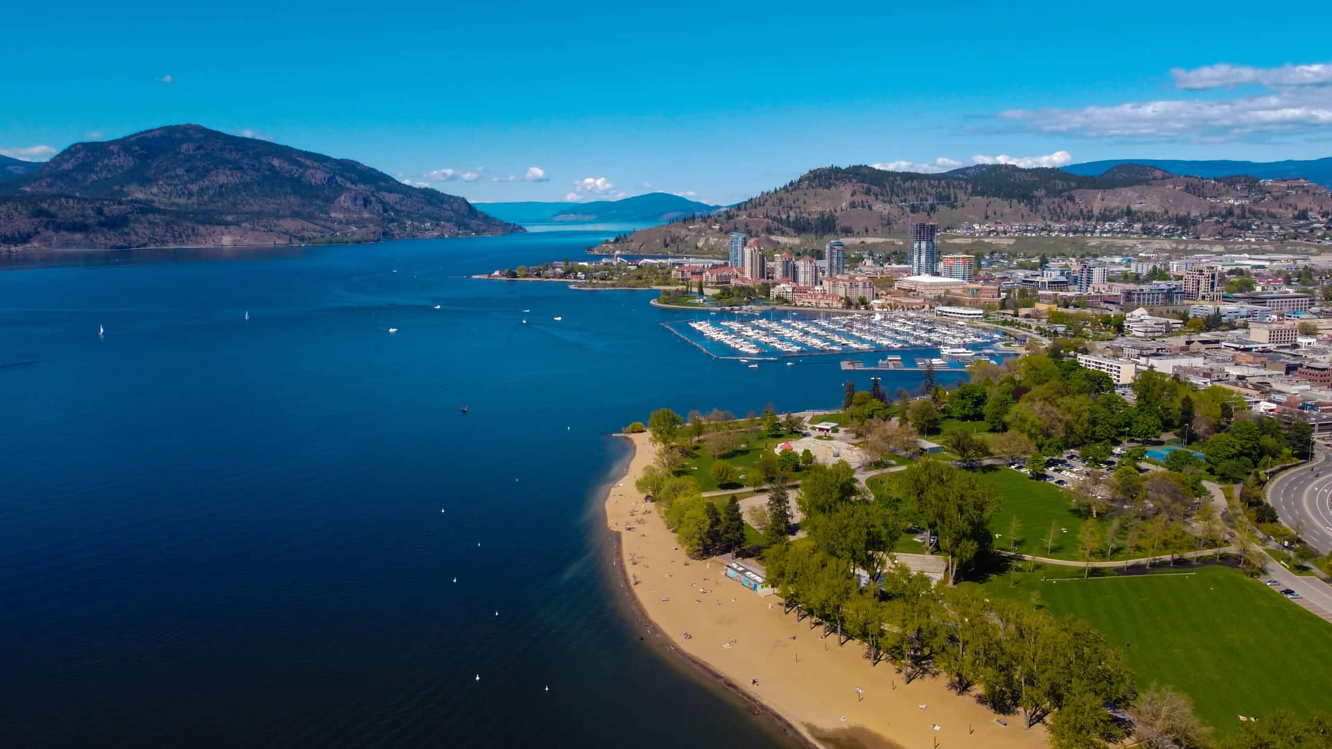 Aerial view of a coastal city with blue water, a sandy beach, greenery, and a marina full of boats. Hills and mountains are visible in the background under a clear blue sky, offering a stunning glimpse of Okanagan's natural beauty—a perfect blend for BC Parks enthusiasts looking to make campsite bookings.