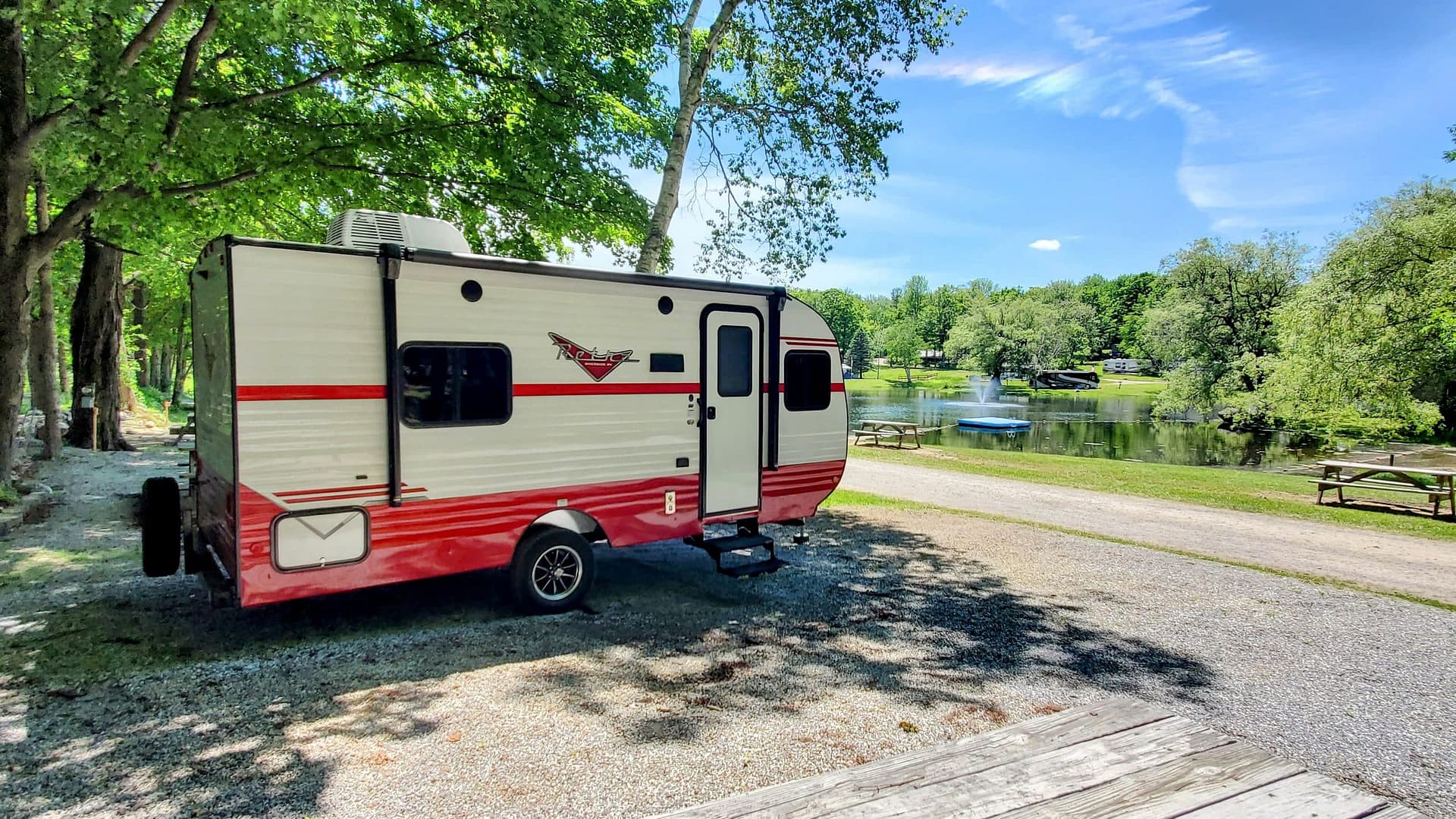 A red and white camper, reminiscent of an auto draft in a travel magazine, is parked on a gravel path near a serene lake. Picnic tables and trees stretch out under the bright, sunny blue sky.