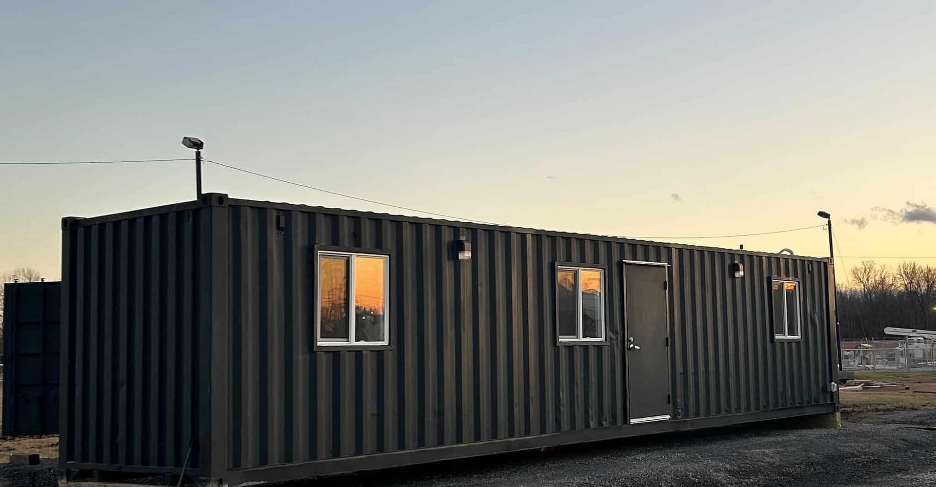 An expansion of ekō Solutions' innovative glamping concept, this black container house sits on a dirt lot.