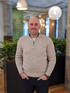 A bald man in a sweater standing in an office discussing marketing technology strategies for Insider Perks.