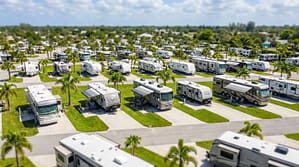 Aerial view of a sprawling RV resort on a sunny day, featuring rows of Class A motorhomes and travel trailers parked on concrete pads with manicured green lawns and palm trees.