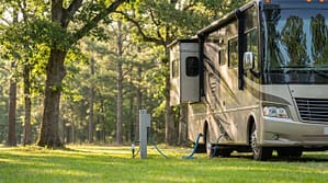 Beige Class A motorhome parked at a grassy campsite with a slide-out extended and connected to a utility pedestal with blue power and water hoses in a wooded area