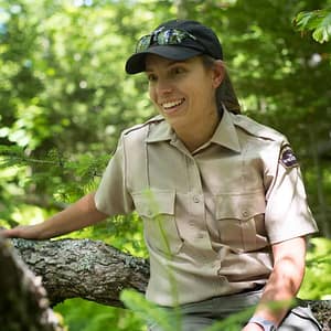 A woman in a uniform from Nova Scotia Parks sitting on a tree in the woods.