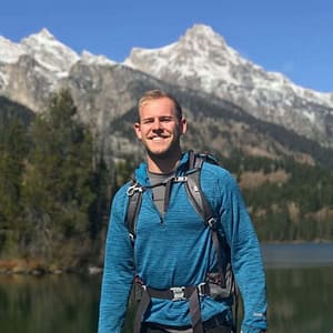 A man standing in front of a lake with mountains in the background during MC Fireside Chats on December 14th, 2022.