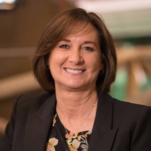 A smiling woman in a black jacket and floral shirt, ready for the MC Fireside Chats on December 21st.