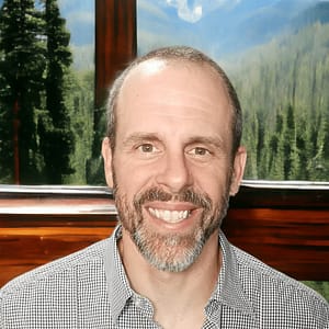 A man smiling in front of a window with mountains in the background during his MC Fireside Chats on August 30th, 2023.