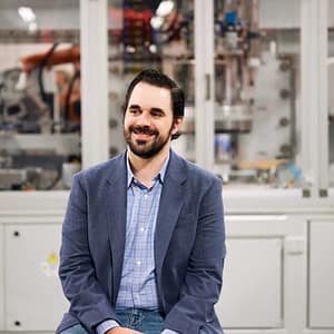 Professional smiling in front of industrial automation equipment during the MC Fireside Chats on March 27th, 2024.