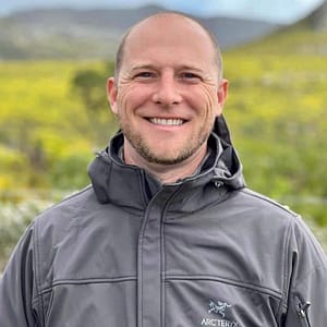 Smiling man wearing a gray jacket standing in front of a field with yellow flowers and a mountain in the auto draft background.