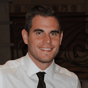 A man with short dark hair and a trimmed beard, wearing a white shirt and black tie, smiles at the camera in an indoor setting, during the May 15th recording of MC Fireside Chats in 2024.