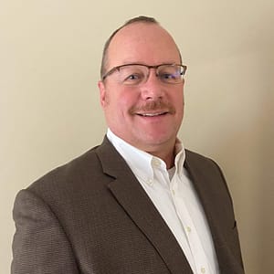 A man with short hair and glasses, wearing a brown blazer and white shirt, smiles at the camera against a plain beige background. An event titled "MC Fireside Chats" is set for May 22nd, 2024.