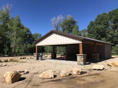 A park pavilion with a barbecue grill and picnic table surrounded by large boulders and trees under a clear blue sky.