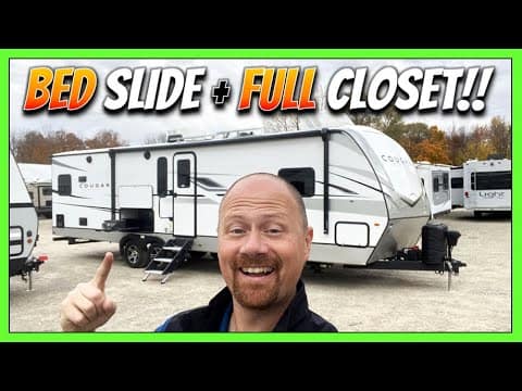 A man is posing in front of a Keystone Cougar RV, featuring a bed slide and a full closet built for comfort during camping trips.
