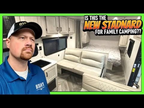 A man is standing in front of an East to West travel trailer with the words, "Is this the standard for family camping?