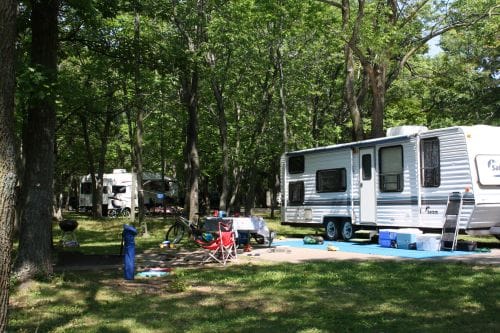 A Brown County Bay Shore Campground scene with a parked RV, scattered camping chairs, a grill, and trees surrounding the area.