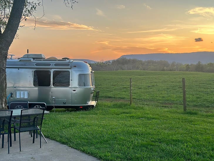 A vintage airstream trailer parked in a grassy field at sunset, with hills in the background and a table and chairs in the auto draft.