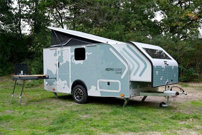 The Aero Cabin One, a small camper trailer with a pop-up roof, is parked in a grassy area for an ultimate camping experience.