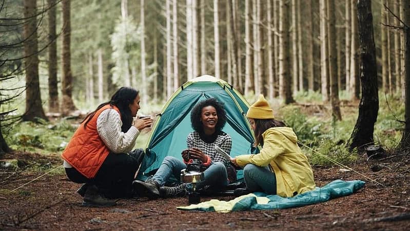 Three melanated women sitting around a tent in the woods during a diverse campout.