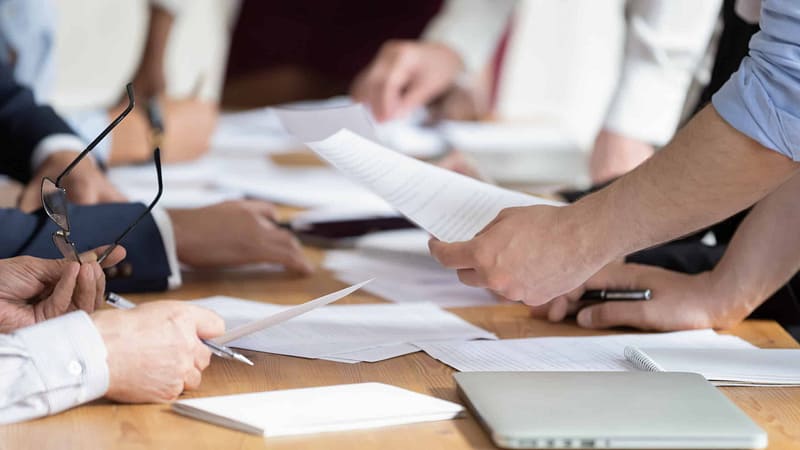 A business meeting in progress with a closeup of participants' hands exchanging auto draft documents over a table.