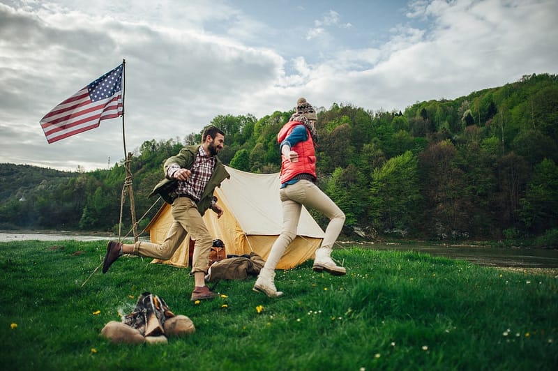 A man and a woman jumping in front of a tent adorned with an American flag, excitedly celebrating the start of Camping Season.