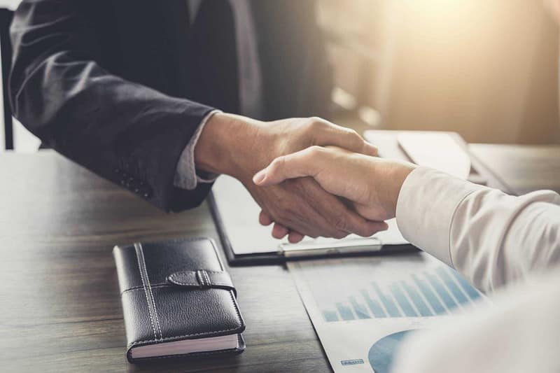 Two business people from PGW Auto Glass Business shaking hands at a desk, signifying the successful transfer of ownership to LKQ Corporation.