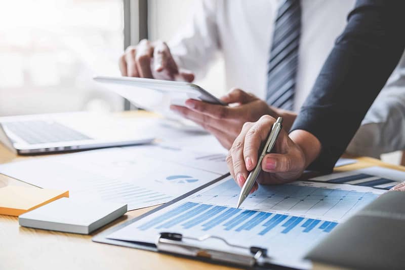 Two business professionals analyzing financial charts and data on a digital tablet and Auto Draft documents at a desk.