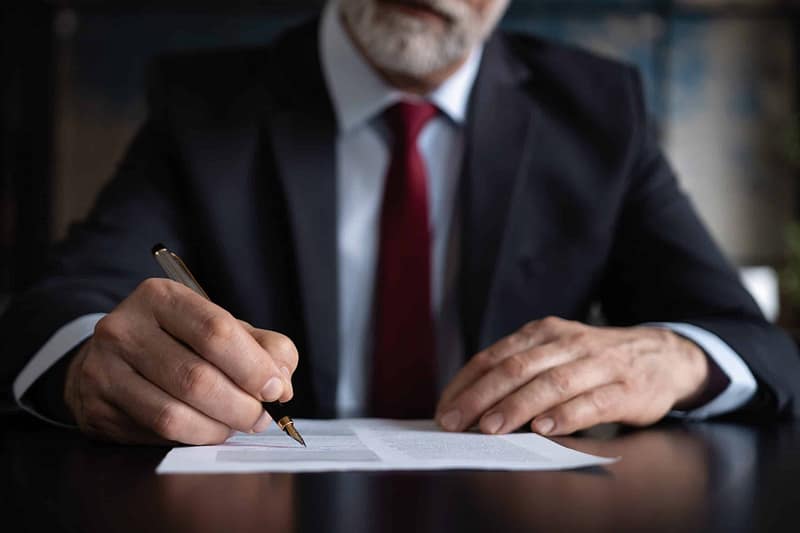 A senior businessman in a suit and red tie signing a USDA document at a desk.
