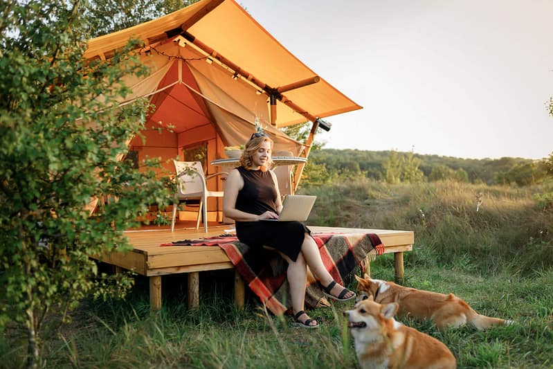 A woman works on a laptop on a porch of a cabin in a field, accompanied by two dogs, during sunset as she manages bookings for Campspot.