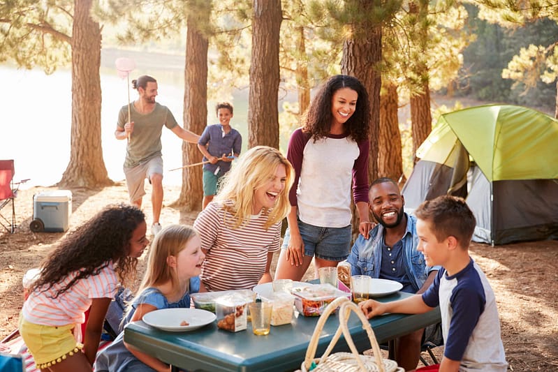 A group of adults and children enjoy a meal at a picnic table in a forested campsite within Mabank's Chuck's RV Resort, with tents in the background. One person in the background holds a fishing net.