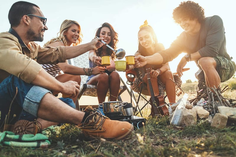 A group of young adults sitting outdoors, laughing and sharing food around a small campfire at Starved Rock Campground during sunset.